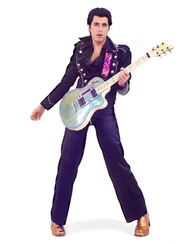 Ein Mann im Elvis-Kostüm posiert mit einer glitzernden Gitarre vor einem weißen Hintergrund.