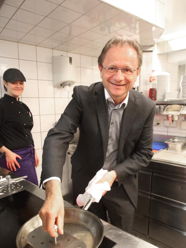 Ein Mann im Anzug hantiert mit einer Pfanne in einer Küche, während eine Köchin im Hintergrund steht.