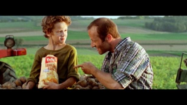 Ein Junge hält eine Tüte Kelly's Chips, während ein Mann auf ihn zeigt; beide sind auf einem Feld.
