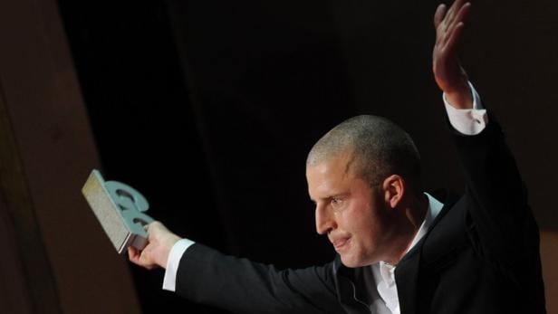 Ein Mann im Anzug hält eine Auszeichnung in der Hand und hebt den Arm.