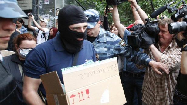 Ein Mann mit Sturmhaube trägt eine beschriftete Kiste, umringt von Polizisten und Kameraleuten.