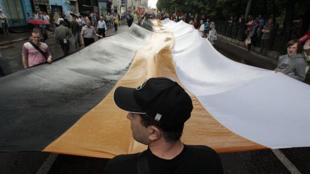 Menschen tragen eine riesige Flagge in den Farben Schwarz, Gold und Weiß durch eine Stadt.