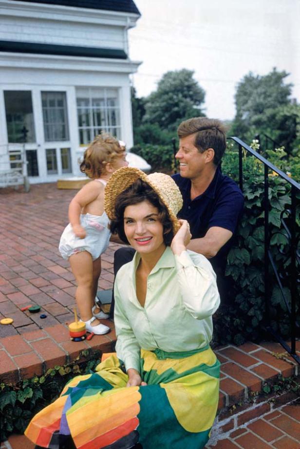 Jacqueline Kennedy sitzt mit Hut auf einer Treppe, während John F. Kennedy und ein Kind im Hintergrund stehen.