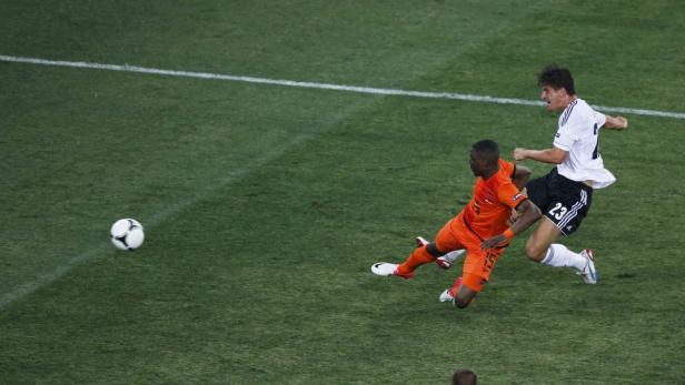 Ein Fußballspieler in Weiß schießt den Ball, während ein Gegenspieler in Orange zu Fall kommt.