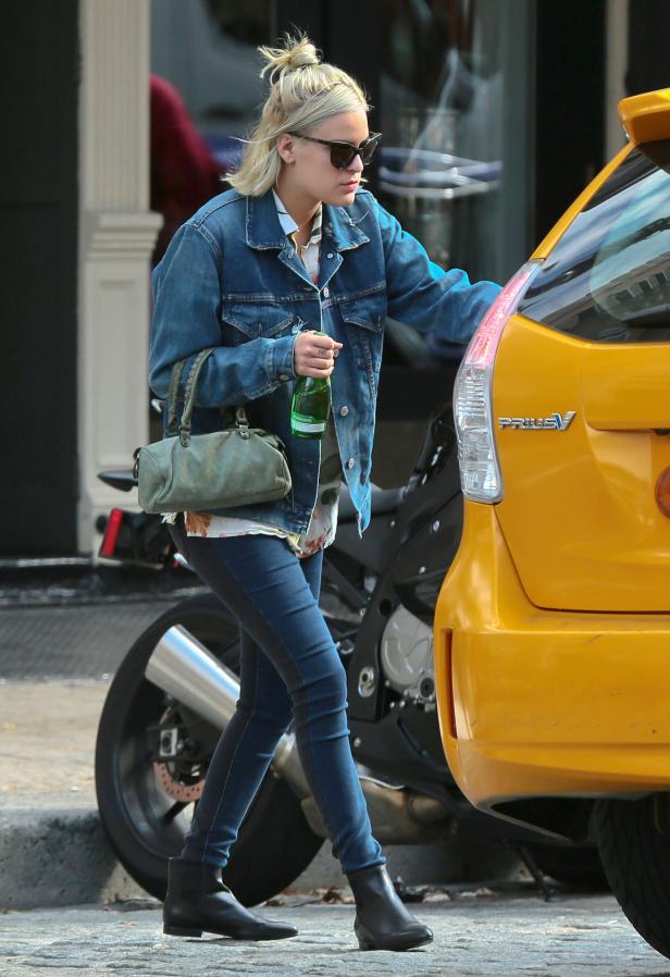 Eine Frau mit Sonnenbrille, Jeansjacke und Handtasche steht neben einem gelben Auto.