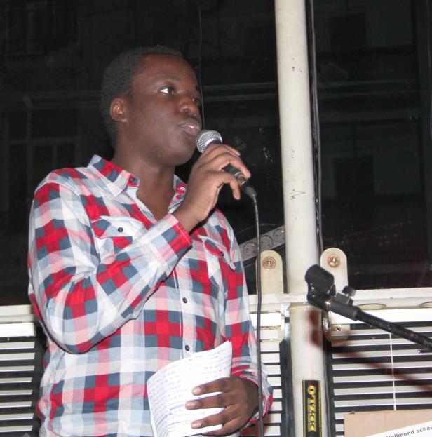 Ein junger Mann mit kariertem Hemd spricht in ein Mikrofon und hält Notizen in der Hand.