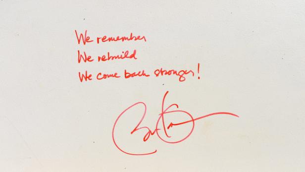 Ein roter Schriftzug auf einer weißen Oberfläche lautet: „We remember, We rebuild, We come back stronger!“.