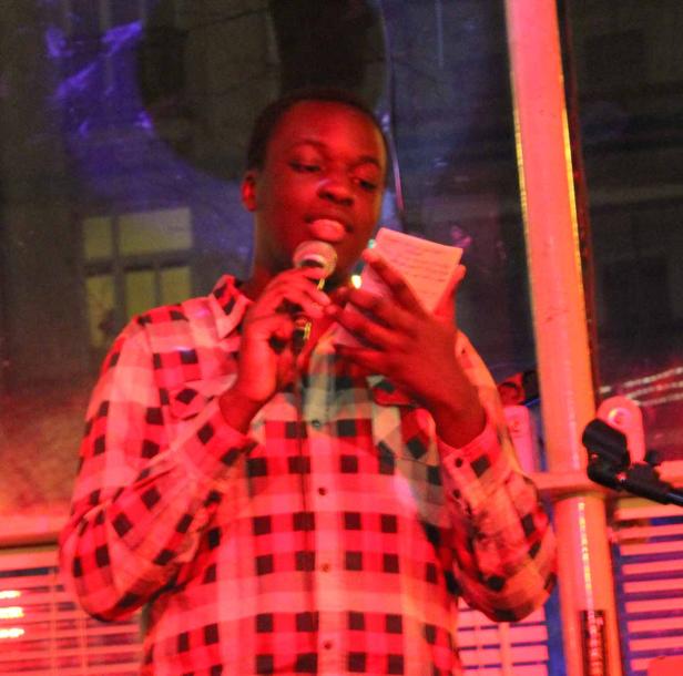 Ein junger Mann mit kariertem Hemd spricht in ein Mikrofon und hält Notizen in der Hand.