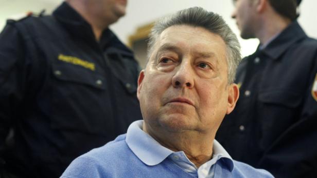 Ein älterer Mann in einem hellblauen Pullover, umgeben von Sicherheitsbeamten.