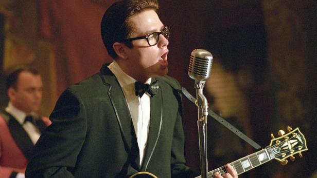 Ein Mann mit Brille und Anzug singt mit Gitarre in ein Mikrofon.
