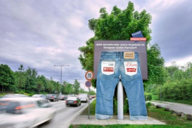 Eine Jeans-Werbetafel für das Designer Outlet Parndorf steht an einer Straße.