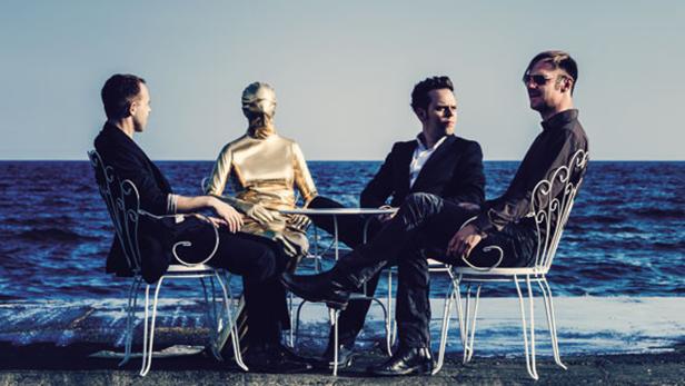 Vier Personen, darunter eine goldene Schaufensterpuppe, sitzen an einem Tisch am Meer.