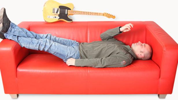 Ein Mann liegt auf einem roten Sofa, über ihm eine gelbe Gitarre.