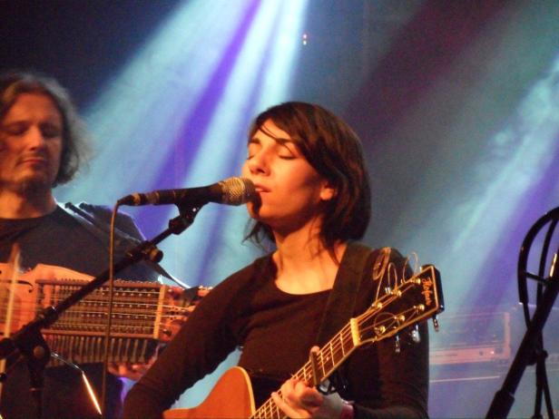 Eine Frau singt und spielt Gitarre auf einer Bühne, während ein Mann ein anderes Instrument spielt.