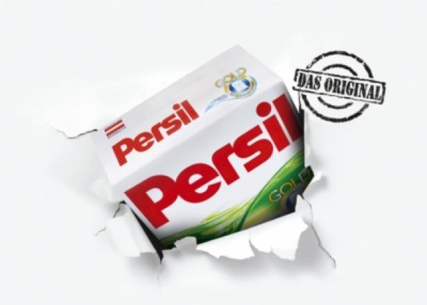 Eine Packung Persil Gold Waschmittel reißt durch eine weiße Papieroberfläche.