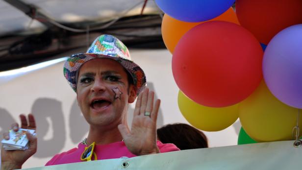 Ein Mann mit Make-up, Hut und bunten Luftballons winkt.