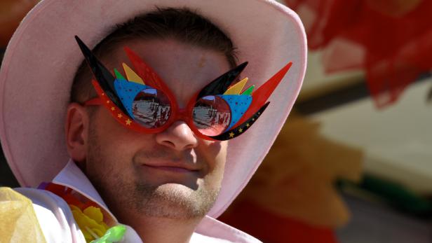 Ein Mann mit rosa Hut und ausgefallener, bunter Brille lächelt.