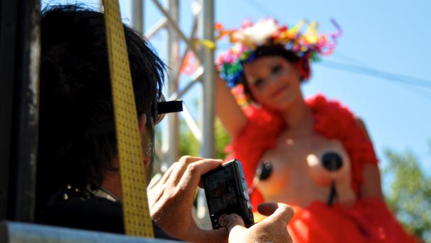 Ein Mann fotografiert eine Frau mit Blumenkranz und roter Boa bei einer Parade.