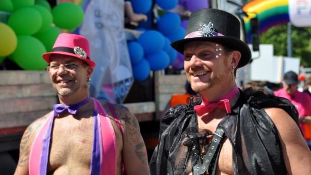 Zwei Männer mit Hüten und Fliegen auf einer Parade, im Hintergrund Ballons und eine Regenbogenfahne.