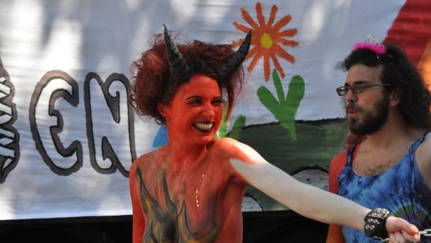 Eine Frau mit roter Körperbemalung und Hörnern lächelt neben einer Person mit Tiara vor einem bemalten Banner.
