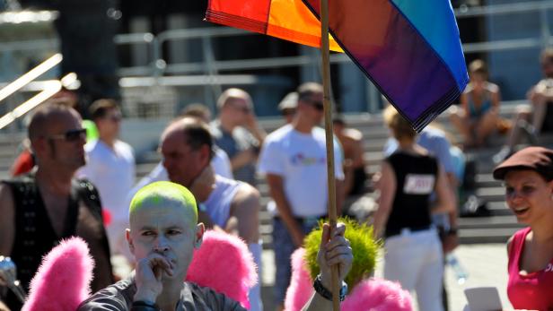 Ein Mann mit grünen Haaren und rosa Flügeln hält eine Regenbogenflagge in einer Menschenmenge.