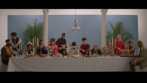 Eine Gruppe von Menschen sitzt an einem langen Tisch mit Speisen und Getränken vor einem blauen Hintergrund.