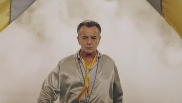 Ein Mann mit ernstem Blick trägt eine goldene Jacke und ein gelbes Halstuch.