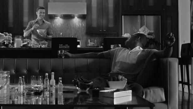 Ein Mann entspannt auf einem Sofa, während ein anderer in der Küche eine Schüssel hält.