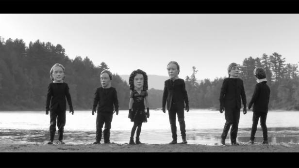 Mehrere Personen mit übergroßen, puppenartigen Köpfen stehen an einem Seeufer.