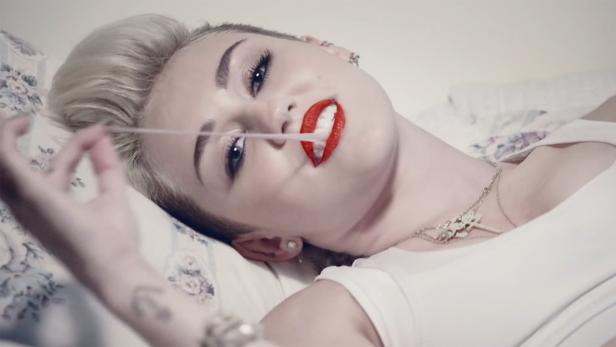 Miley Cyrus liegt auf einem floralen Kissen und spielt mit einem Gummiband.