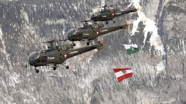 Drei österreichische Militärhubschrauber fliegen vor einer verschneiten Berglandschaft mit Flaggen.