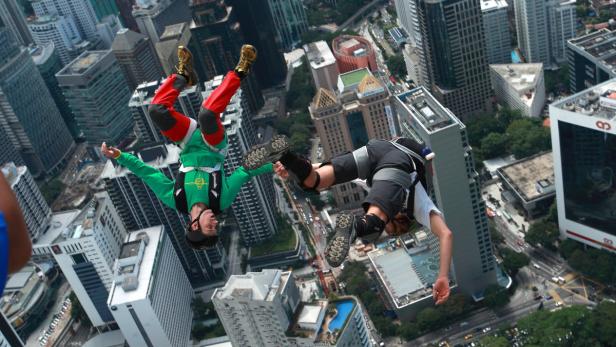 Zwei Fallschirmspringer stürzen sich vor einer Stadtkulisse in die Tiefe.