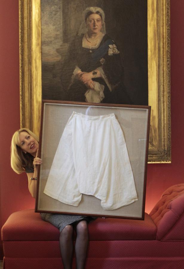 Eine Frau präsentiert ein Paar ausgestellte, gerahmte Unterhosen vor einem Porträt von Königin Victoria.