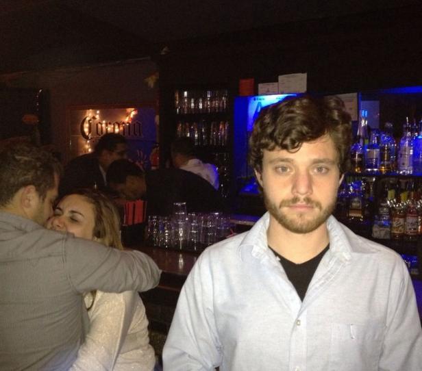 Ein Mann steht in einer Bar, im Hintergrund ein Paar beim Küssen.