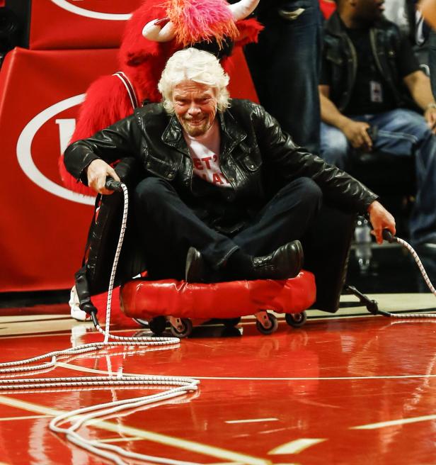 Richard Branson sitzt auf einem roten Rollbrett auf einem Basketballfeld.