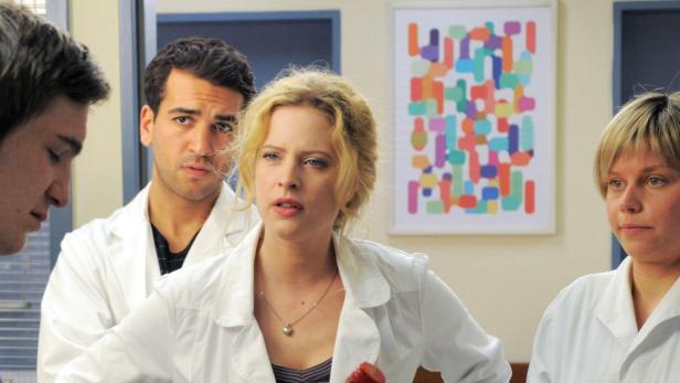 Vier Personen in weißen Arztkitteln stehen in einem Raum mit einem bunten Bild an der Wand.