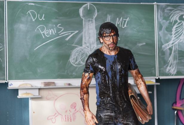 Ein mit Schokolade beschmierter Mann steht vor einer Tafel mit obszönen Zeichnungen.