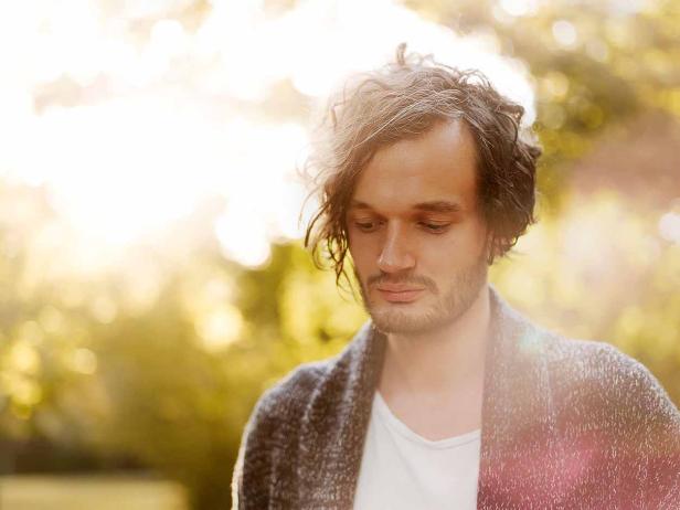 Ein Mann mit lockigem Haar und Bart trägt eine Strickjacke.