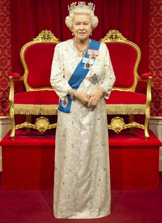 Eine Wachsfigur von Königin Elisabeth II. mit Krone und blauem Ordensband vor einem roten Thron.