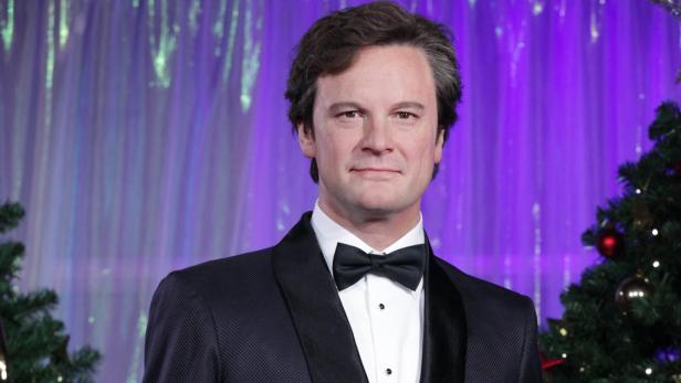 Colin Firth in einem schwarzen Smoking mit Fliege vor einem Weihnachtsbaum.