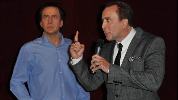 Nicolas Cage spricht neben seiner Wachsfigur in einem Mikrofon.