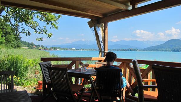 Eine Person sitzt auf einer Terrasse mit Blick auf einen See und die Berge.