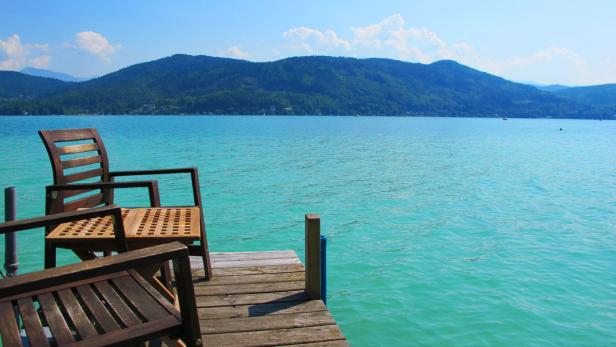 Zwei Holzstühle stehen auf einem Steg mit Blick auf einen See und bewaldete Hügel.