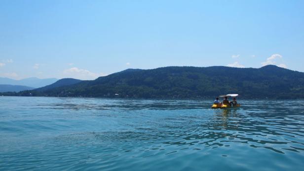 Ein gelbes Tretboot mit Personen fährt auf einem See vor bewaldeten Hügeln.