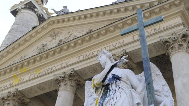 Eine mit Farbe beschmierte Engelsstatue vor einer Kirche mit einem Kreuz im Vordergrund.