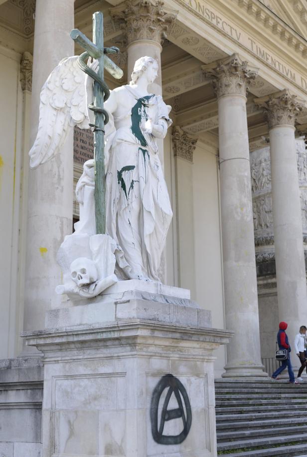 Eine Statue eines Engels vor einem Gebäude, beschmiert mit Farbe und einem Anarchie-Symbol.