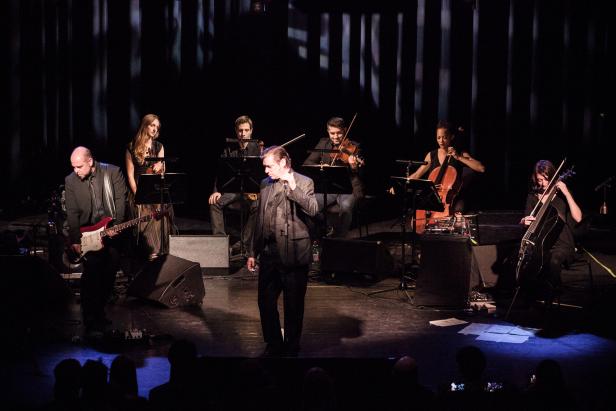 Ein Sänger steht mit seiner Band auf einer dunklen Bühne.
