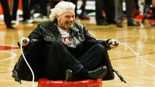 Richard Branson sitzt auf einem roten Kissen und lächelt.