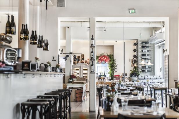 Innenansicht eines hellen Restaurants mit Tischen, Stühlen und dekorativen Weinflaschen als Lampen.