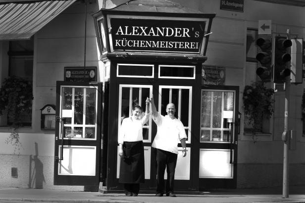 Zwei Personen stehen vor „Alexander's Küchenmeisterei“ und heben die Arme.